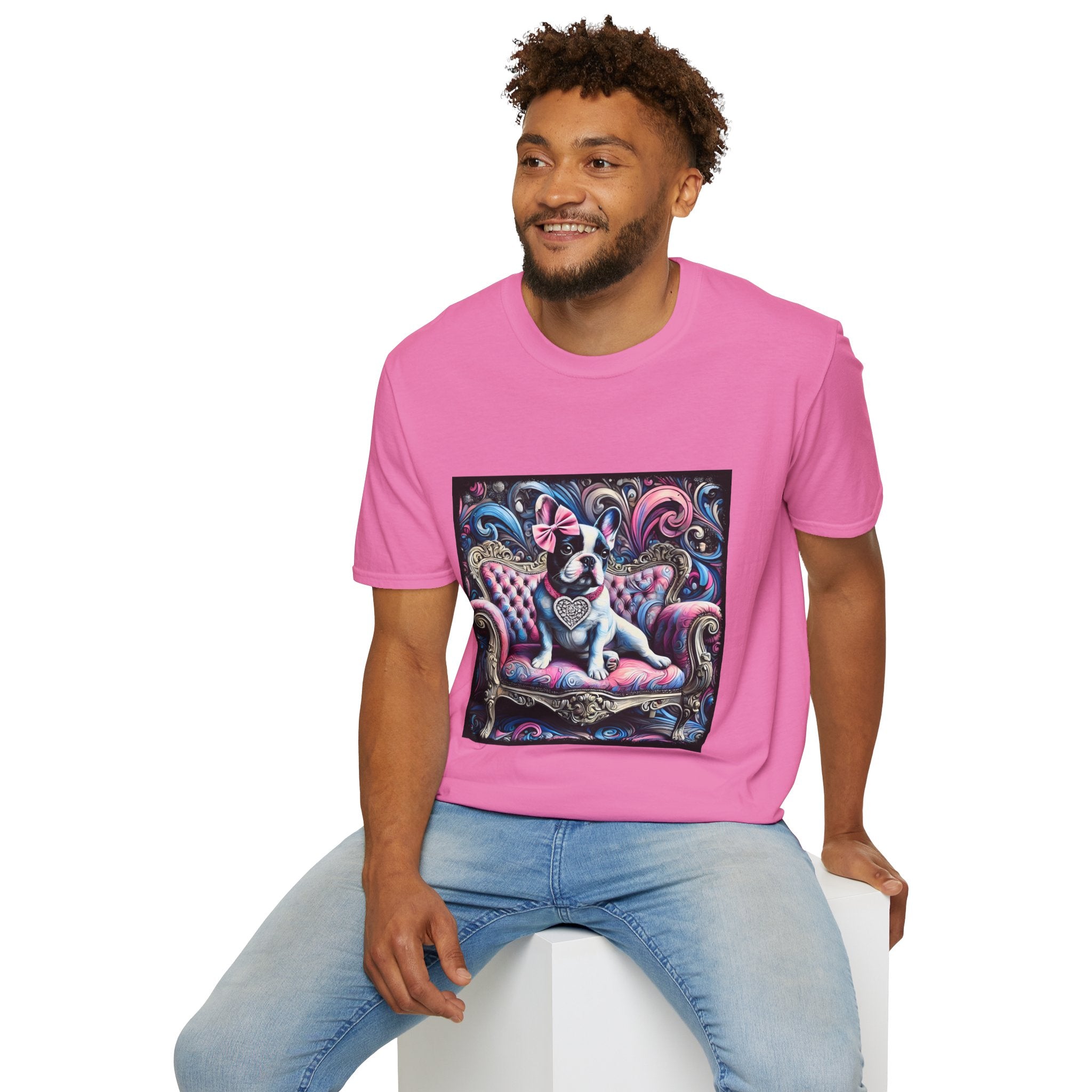 Male model in pink dog t-shirt featuring a black and white French Bulldog with a pink bow, seated on a tufted pink and gold settee against a swirling blue, pink, and purple expressionist-style background.